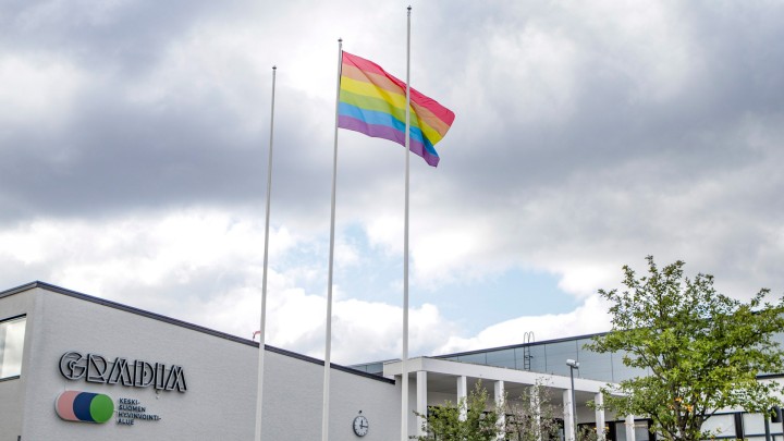 Gradian rakennus, jonka edessä on kolme lipputankoa; keskimmäisessä liehuu sateenkaarilippu. Taustalla pilvinen taivas, toinen rakennus ja puita.